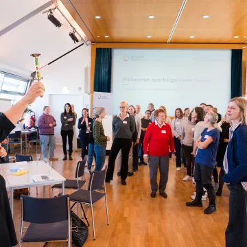 © Stefanie J. Steindl/Gesundheit Österreich GmbH Teilnehmende des Bürger:innenforums zur Stärkung der Patient:innen- und Bevölkerungsbeteiligung im Gesundheitswesen in Wien, organisiert von der Gesundheit Österreich GmbH. Eine Moderatorin eröffnet die Veranstaltung mit einem symbolischen Signalhorn.