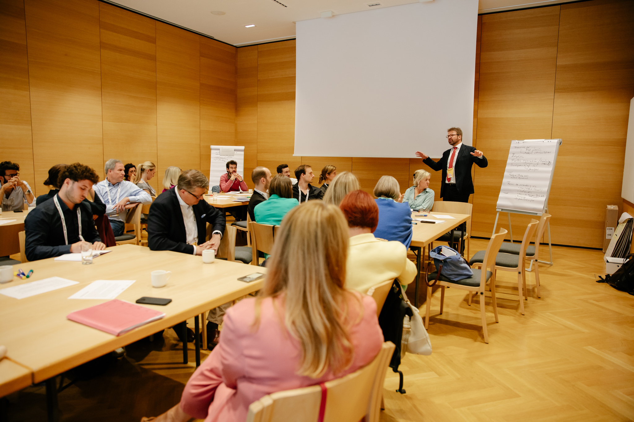 Teilnehmende einer EHDS-Worksession sitzen in einem Seminarraum an mehreren Tischen. Eine Person leitet die Diskussion vorne im Raum und erklärt Inhalte an einem Flipchart. Die Gruppe hört zu, macht Notizen und arbeitet gemeinsam an Fragestellungen rund um die Nutzung von Gesundheitsdaten.
