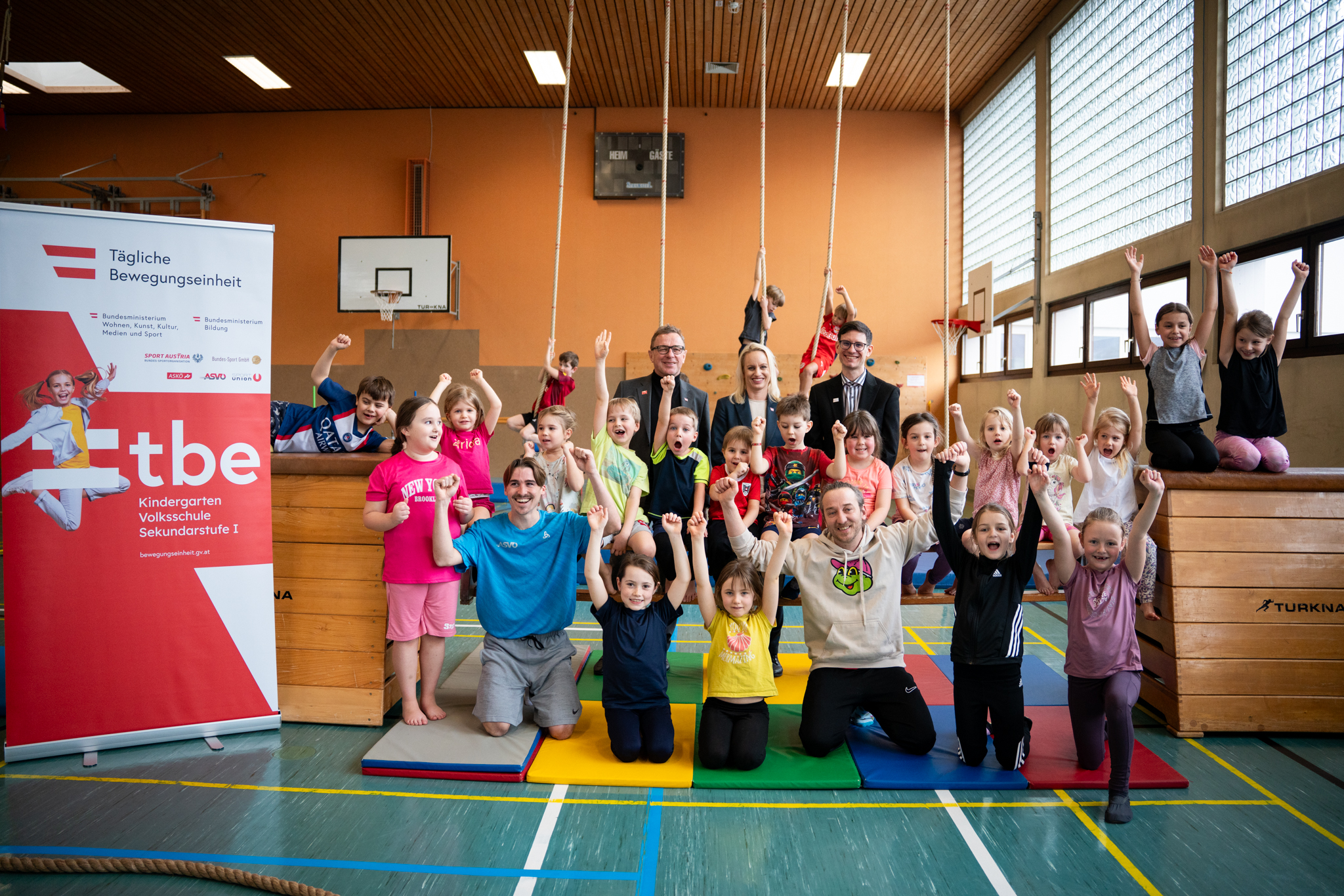 Gruppenfoto in einer Turnhalle: Kinder und Erwachsene jubeln vor einem Roll-up zur „Täglichen Bewegungseinheit“.
