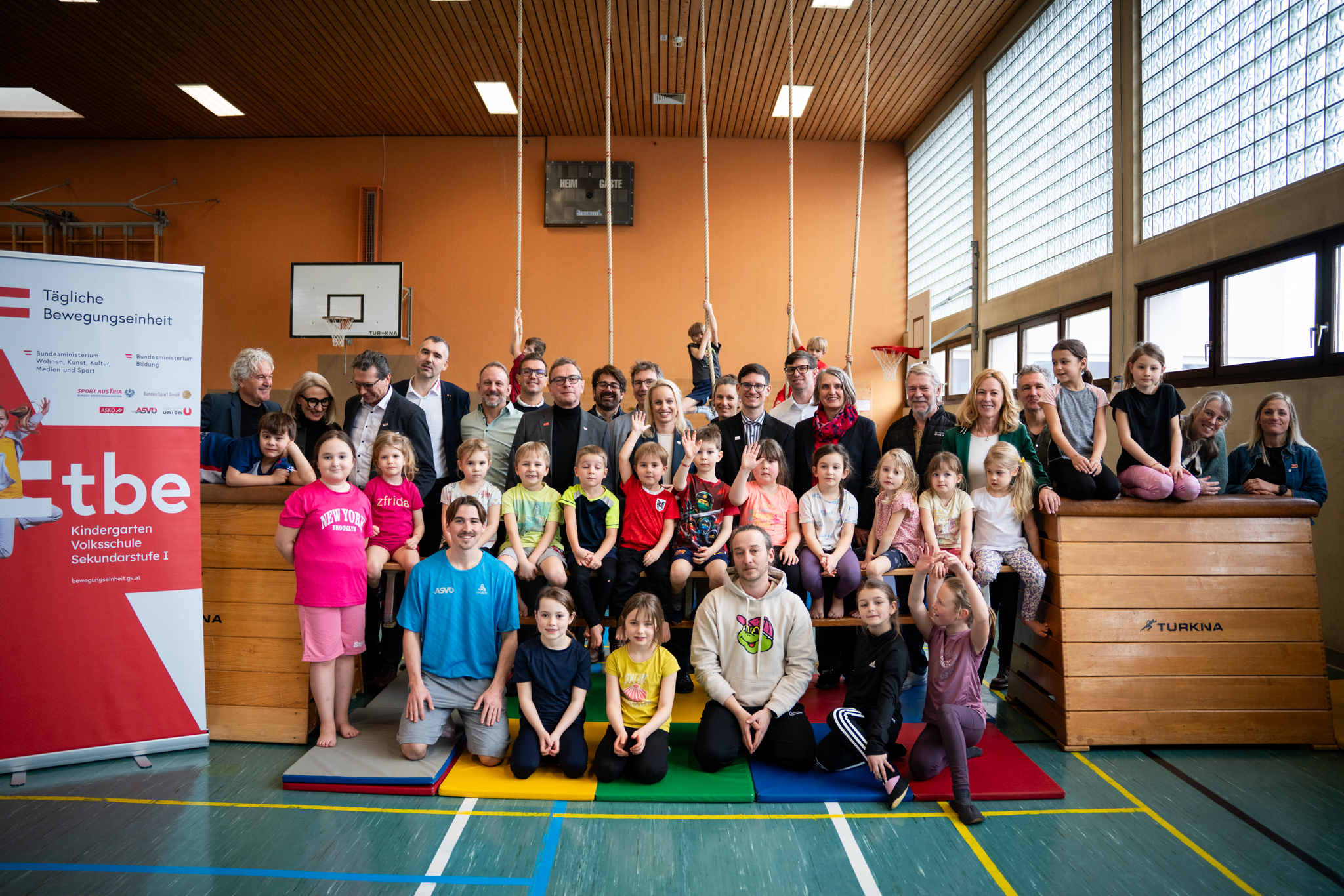 Großes Gruppenfoto in einer Turnhalle: Kinder und Erwachsene sitzen und stehen vor einem Roll-up zur „Täglichen Bewegungseinheit“.