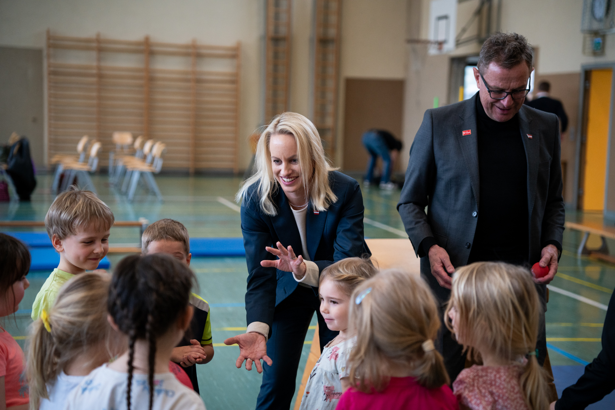 Sport-Staatssekretärin Michaela Schmidt und Ralf Rangnick sprechen in einer Turnhalle mit einer Gruppe von Kindern.