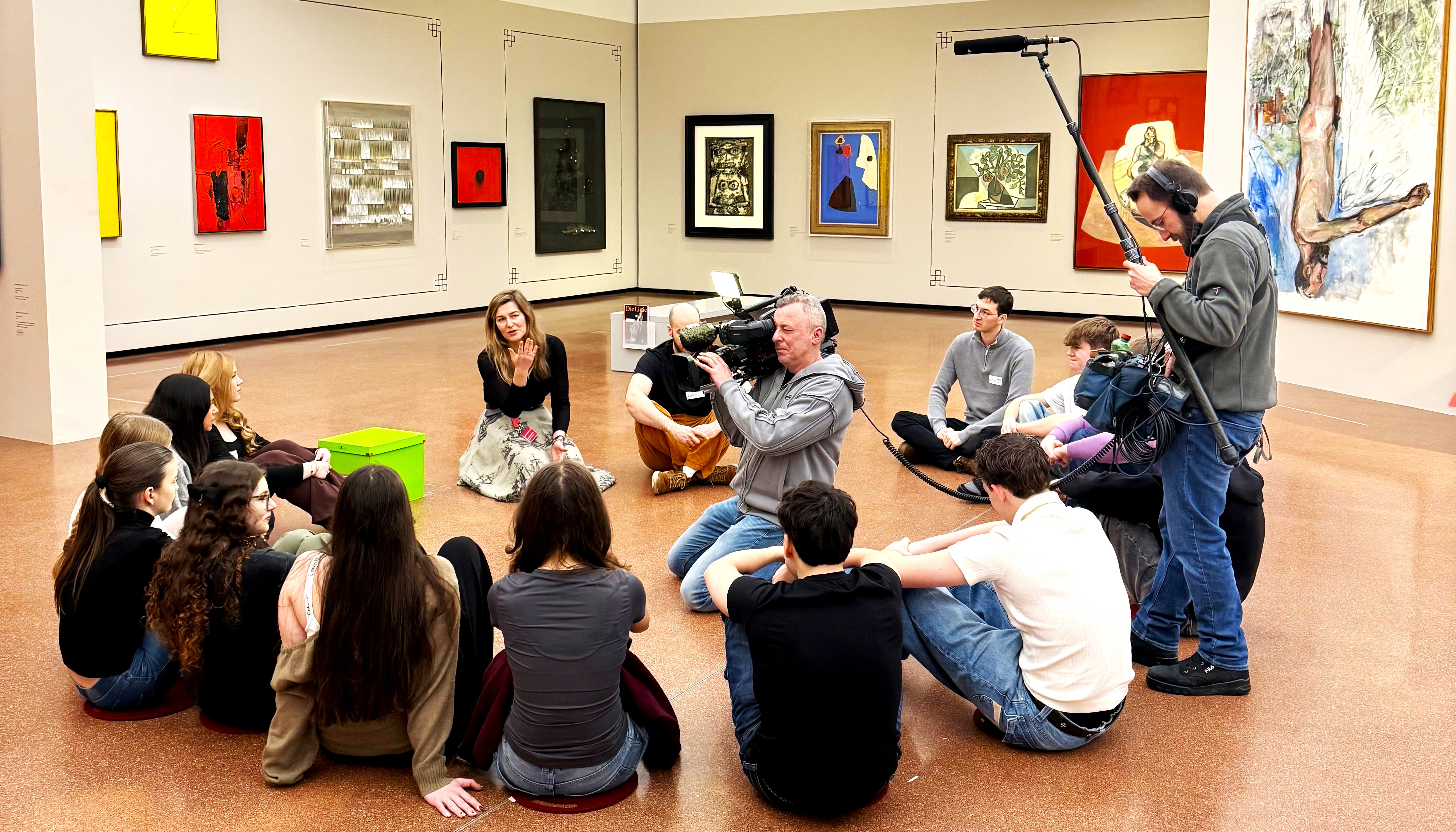 Jugendgruppe sitzt im Kreis im Museumsraum; ein Kamerateam filmt und nimmt Ton auf, während eine Vermittlerin mit den Jugendlichen spricht.