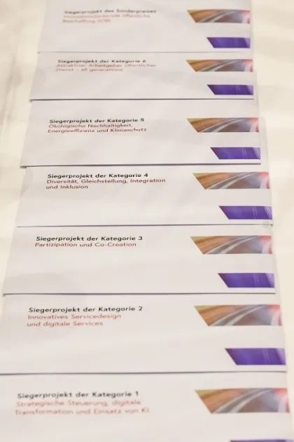 Nahaufnahme von sieben Broschüren mit der Aufschrift ‚Siegerprojekt‘ der jeweiligen Kategorien des Österreichischen Verwaltungspreises 2025 – von KI und digitalen Services bis zu Diversität, Nachhaltigkeit und Co-Creation.